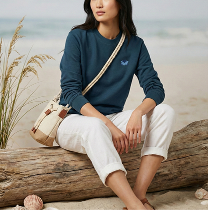 Woman sitting on a log by the beach wearing a blue sweater and white pants.